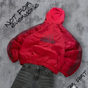Vintage Adidas ”Varm” Vindjacka - Säljer en cool röd varm vindjacka från Adidas med svart/grå detaljer och klassiska ränder längs ärmarna. Jackan har en dragkedja framtill och en huva för extra skydd. Perfekt för en sportig look! Storlek L