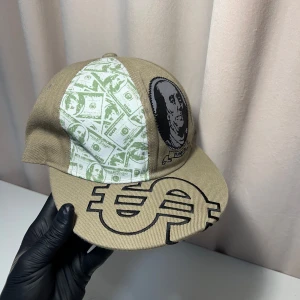 Beige keps med dollartryck - Cool beige keps med unikt dollartryck och porträtt av Benjamin Franklin. Kepsen har en platt skärm med ett stort dollartecken i svart. Perfekt för att sticka ut med en stilfull look.