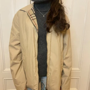 Burberry jacka - Säljer en snygg beige vändbar jacka från Burberry. Ena sidan är enfärgad beige medan den andra har det klassiska Burberry-rutiga mönstret. Jackan har långa ärmar och en dragkedja framtill. Perfekt för att variera din stil!