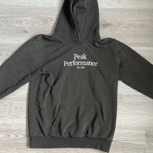  hoodie från Peak Performance - Säljer en  hoodie från Peak Performance med vit logga på bröstet. Tröjan har en klassisk känguruficka och ribbade muddar. Perfekt för en avslappnad stil. Tyvär några som slitningar men det syns knappast