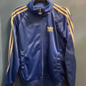 Blå sporttröja från Adidas - Säljer en klassisk blå träningsjacka från Adidas(inte helt hundra om den är äkta) med vita ränder längs ärmarna och dragkedja framtill. Jackan har ribbade muddar och det ikoniska Adidas-loggan på bröstet. Storlek L (liten), pris kan diskuteras!