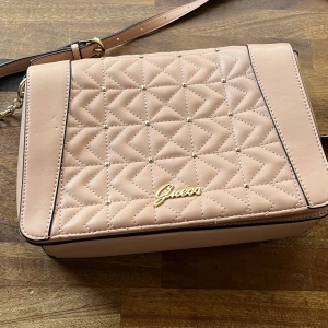 Rosa quiltad axelväska från Guess - Snygg rosa axelväska från Guess med quiltad design och guldfärgad logga framtill. Väskan har en justerbar axelrem och ett mönstrat innerfoder. Perfekt för att ge din outfit en stilren touch.