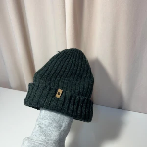 Fjällräven mössa - Säljer en stilren grön ribbad mössa från Fjällräven. Mössan har en enkel design med en liten etikett framtill. Perfekt för att hålla värmen under kyliga dagar.