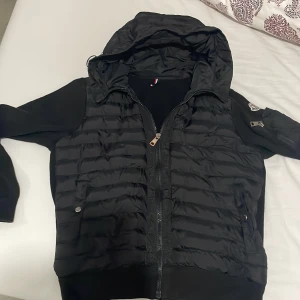 Moncler cardigan - Säljer en svart jacka från Moncler med huva och dragkedja. Jackan har långa ärmar och ett quiltat mönster på framsidan. Den är tillverkad i en kombination av material för en stilren look. Perfekt för kyligare dagar. Pappret på insidan har gått av men de är lätt o sy fast den igen jag är 175 jackan är perfekt storlek 1/s