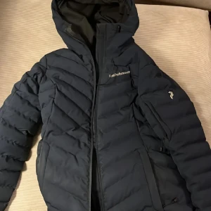 Mörkblå dunjacka från Peak Performance - Snygg och varm mörkblå dunjacka från Peak Performance med huva och dragkedja.