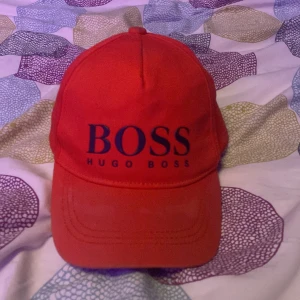 Röd keps från Hugo Boss - Snygg röd keps från Hugo Boss med justerbart spänne baktill. Kepsen har en klassisk design med märkeslogga framtill och på spännet. Perfekt för en stilren look. Lite smutsig men går att tvätta bort! Ganska mycket använd, ock äkta!. Tveka inte på att höra av dig!