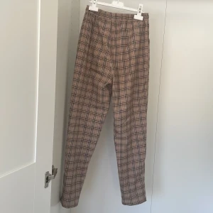 Rutiga byxor från Next - Snygga rutiga byxor från Next i beige och bruna toner. Byxorna har en elastisk midja för extra komfort och är tillverkade i en mjuk polyesterblandning. Perfekta för en stilren look.