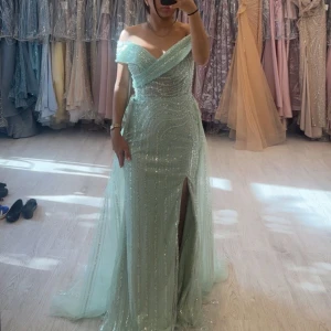 Mintgrön glittrig långklänning - Elegant mintgrön långklänning med glittrande detaljer och en slits. Klänningen har en offshoulder-design och är perfekt för festliga tillfällen såsom bal eller bröllop! Använd en gång, originalpris 8000. Kommer med tillhörande dustbag. Pris kan diskuteras vid snabb affär