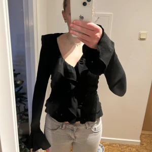 Svart blus med volangdetaljer - Säljer en svart blus med volangdetaljer och långa ärmar. Blusen har en v-ringad design. Perfekt för en stilren look.