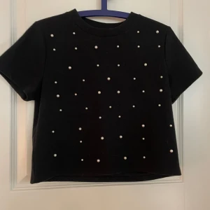 Svart kortärmad topp med pärlor - Snygg svart topp med korta ärmar och dekorativa pärlor på framsidan. Perfekt för att ge en stilren look med en touch av elegans.