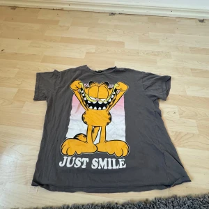 Grå t-shirt med Garfield-tryck från FB Sister - Säljer en grå t-shirt från FB Sister med ett stort Garfield-tryck och texten 'Just Smile' på framsidan. T-shirten är kortärmad och har en avslappnad passform. Perfekt för en avslappnad stil med en humoristisk touch!