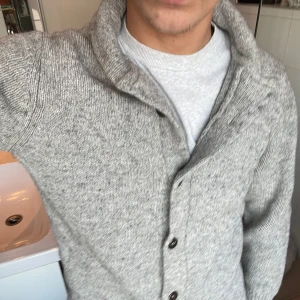 Grå stickad kofta  - Säljer en stilren grå stickad kofta. Koftan har en klassisk design med knappar framtill och en hög krage. Perfekt för kyligare dagar och ger en snygg look till vardagsoutfiten.