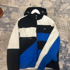 Flerfärgad vindjacka från Lacoste - Säljer en snygg flerfärgad vindjacka från Lacoste i svart, blått och vitt. Jackan har en dragkedja framtill och en huva. Perfekt för blåsiga dagar och har en sportig look.