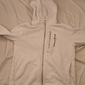 Vit huvtröja från Sail Racing - Snygg vit huvtröja från Sail Racing med dragkedja framtill och en liten bröstficka med dragkedja. Perfekt för en stilren look. Huvan ger extra komfort och värme.vi kan diskutera priset