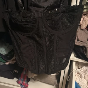 Svart korsett med spetsdetaljer - Snygg svart korsett med spetsdetaljer och justerbara axelband. Den har hyskknäppning bak och är perfekt för en elegant look. Passar bra till både fest och vardag.