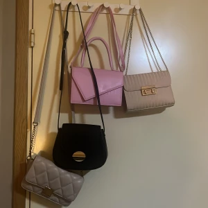 Fyra eleganta väskor i olika färger - Säljer fyra snygga väskor: en rosa handväska med krokodilmönster, en beige axelväska med kedja och gulddetaljer, en svart axelväska med rund gulddetalj, och en quiltad beige axelväska med silverdetalj. Perfekta för att komplettera din outfit! Du kan köpa en av dom om du vill behövs inte köpa alla och den rosa är helt ny aldrig använt 80kr för den rosa väskan 
