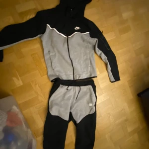Nike tech tracksuit - Säljer en grå och svart tracksuit från Nike med dragkedja och huva. Jackan har långa ärmar och en sportig design med Nike-logga på bröstet. Perfekt för träning eller en avslappnad stil. 🖤pris kan diskuteras
