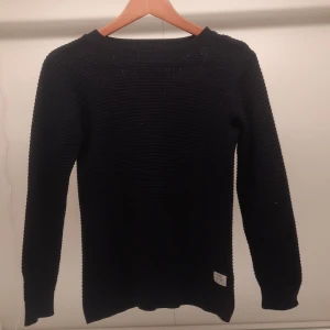 Svart stickad tröja från 157 - Säljer en svart stickad tröja från 157. Tröjan har en ribbad struktur och långa ärmar, perfekt för kyligare dagar. Den är enkel och tidlös i designen, vilket gör den lätt att matcha med olika outfits.