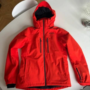Röd skidjacka från EQPE - Snygg röd skidjacka från EQPE med huva och dragkedja. Jackan har flera praktiska fickor med dragkedjor och är perfekt för blåsiga dagar. Den har en modern design och är både funktionell och stilren. Endast använd 3 veckor.