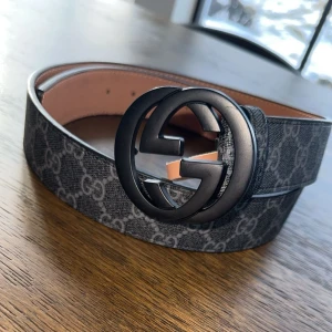 Svart bälte  - Snyggt svart bälte f med ett klassiskt monogrammönster. Bältet har ett stort, runt spänne i svart. Perfekt för att ge en lyxig touch till din outfit.