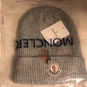 Grå ribbad mössa från Moncler - Snygg grå ribbad mössa från Moncler med broderad logotyp framtill. Perfekt för kyliga dagar och ger en stilren look. Mössan är i en neutral färg som passar till det mesta.