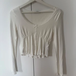 Vit långärmad topp med spetsdetaljer - Vit långärmad topp med spetsdetaljer och liten rosett framtill. Toppen är croppad med volangdetaljer och har en v-ringad design. 