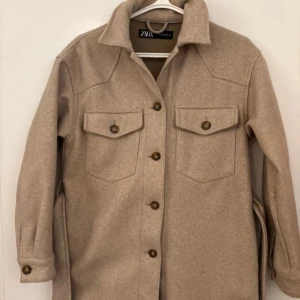 Kappa  från Zara - Snygg beige kappa från  Zara med knappar och två bröstfickor. Jackan har långa ärmar och ett bälte i midjan för en stilren look. Perfekt för lager-på-lager under hösten.
