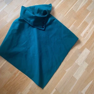 Grön poncho med knappar - Snygg grön poncho med hög krage och tre dekorativa knappar. Perfekt för att hålla sig varm och stilren under kyliga dagar. Den har en enkel och elegant design som passar till många olika outfits. Storlek M 