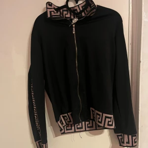 Svart jacka med mönstrade detaljer - Snygg svart jacka med dragkedja och unika mönstrade detaljer i beige längs ärmarna och nederkanten. Perfekt för att ge en stilren look en extra touch. Jackan har långa ärmar och en hög krage.