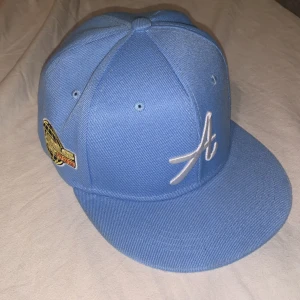 Blå Keps - Snygg blå keps med en vit broderad 'A' på framsidan och en färgglad patch med texten 'Los Angeles Air Sport 1985' på sidan. Perfekt för en casual look.