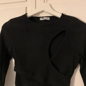 Svart ribbad topp - Snygg svart ribbad topp med lång ärm och en unik cut-out detalj på framsidan. Perfekt för att ge din outfit en edgy touch. Toppen är croppad och passar bra till både jeans och kjol.
