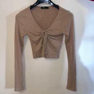 Beige ribbad topp från FB Sister - Säljer en beige ribbad topp från FB Sister. Den har en v-ringad design med knytning framtill och är croppad. Perfekt för en stilren look.