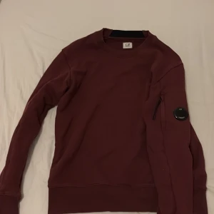 Vinröd tröja från C.P. Company - Säljer en stilren vinröd tröja från C.P. Company. Tröjan har en rund hals och en praktisk ficka med dragkedja på ärmen. Perfekt för en avslappnad look.