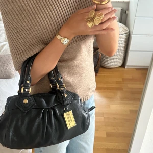 Svart Q Classic Baby Groove väska! - Snygg svart handväska i bra skick från Marc by Marc Jacobs i äkta läder med gulddetaljer och dubbla handtag. Finns lite slitage på fötterna på undersidan, men inget tydligt alls. Inga repor eller hål på väskans läder. Små slitningar finns på några gulddetaljer. Välbevarad vintage modell! Perfekt för att ge en stilren touch till din outfit. Skriv privat för fler bilder. Mått 34x18x12 (undersida). Kan hämtas hemma hos mig i Nacka eller skickas men kan tyvärr inte mötas🤍