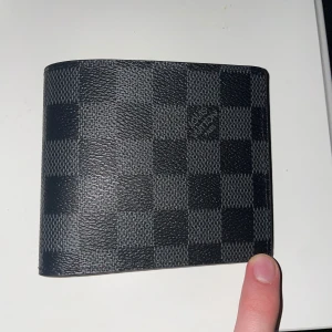 Rutig plånbok från Louis Vuitton - Säljer en stilren svart plånbok från Louis Vuitton med klassiskt rutigt mönster. Plånboken har flera kortfack och en sektion för ID-kort. Perfekt för dina kort och kontanter. Denna är FEJK men väldigt bra gjord och har aldrig användts.  Detta pris gäller självklart inte, kom med prisförslag