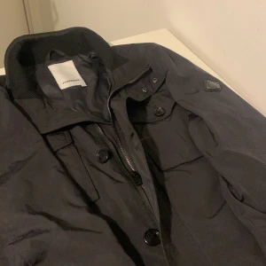 Svart jacka från J.Lindeberg - Snygg svart jacka från J.Lindeberg med dragkedja och knappar. Perfekt för en stilren look. Jackan har en diskret logotypdetalj på ärmen och en klassisk krage. Nypris runt 3000-3500 mitt pris 2499