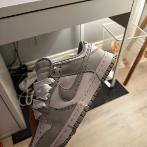 Nike Dunk grå vit  - Snygga Nike Dunk sneakers i en stilren kombination av grått och vitt. Skorna har en klassisk design med snörning och en bekväm sula. Perfekta för en avslappnad stil.
