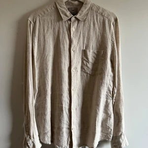 Beige linneskjorta från H&M - Säljer en stilren beige skjorta från H&M i linne. Skjortan har en klassisk krage och knappar framtill. Perfekt för en avslappnad look med en bröstficka som extra detalj. Regular fit för en bekväm passform.