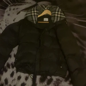 Burberry jacka/väst - Säljer en snygg svart dunjacka från Burberry med avtagbara ärmar. Jackan har en klassisk rutig huva och dragkedja framtill. Perfekt för kyliga dagar när du vill hålla stilen. Storlek L men sitter mer som M är justerbar i storleken också. Kan bytas mot en mindre storlek eller nått annat