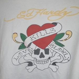Vit hoodie från Ed Hardy - Säljer en vit hoodie från Ed Hardy med ett stort tryck av ett hjärta och en dödskalle på framsidan. Texten 'Love Kills Slowly' är tryckt över motivet. Hoodien har dragkedja och fickor framtill samt justerbar huva med snören.