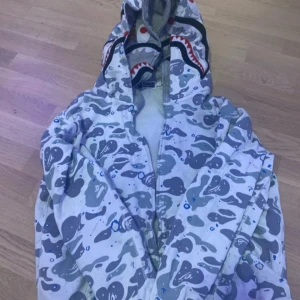 Camouflage hoodie från A Bathing Ape - Säljer en cool hoodie från A Bathing Ape med blått camouflage-mönster och hajdesign på huvan. Den har broderade bokstäver 'WGM' i färgerna röd, blå och orange. Perfekt för dig som vill sticka ut!
