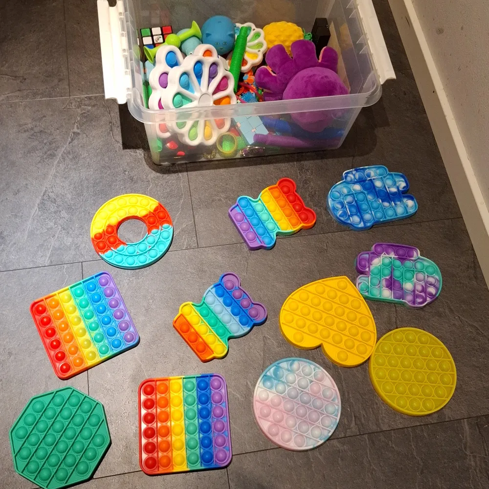 Stor samling av pop it-leksaker i en genomskinlig plastlåda. Innehåller olika former som fyrkantiga, runda och hjärtformade i regnbågsfärger, gult och blått. Perfekt för stressavlastning och sensorisk lek. Inkluderar även en Rubiks kub och andra små fidget-leksaker.. Muu.