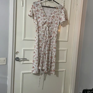 Blommig klänning  - En vit klänning med blommigt mönster. Den har en v-ringad design och korta ärmar. Knytband i midjan för en fin passform. Perfekt för sommaren och andra tillfällen ❤️Klänningen har inte används av mig och är som ny☺️