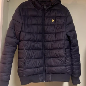 Marin blå pufferjacka från Lyle & Scott - Snygg Marin blå pufferjacka från Lyle & Scott med gul logga på bröstet. Jackan har en dragkedja framtill och en huva för extra skydd mot väder. Perfekt för alla årstider.