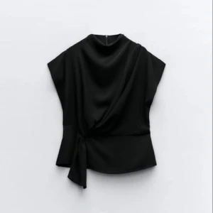 Svart draperad topp - Elegant svart topp med draperad design och knytning i sidan. Toppen har en hög hals och korta ärmar, vilket ger en stilren look. Perfekt för en chic och modern stil.