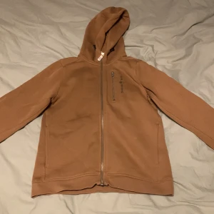 Brun hoodie från Sail Racing - Säljer en brun hoodie från Sail Racing med dragkedja och huva. Den har en stilren design med en liten logotyp på bröstet och en praktisk ficka med dragkedja. Perfekt för en avslappnad stil.