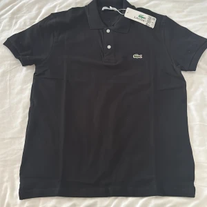 Svart pikétröja från Lacoste - Snygg svart pikétröja från Lacoste med klassisk krage och knappar. Tröjan har det ikoniska krokodilmärket broderat på bröstet. Perfekt för en stilren look.