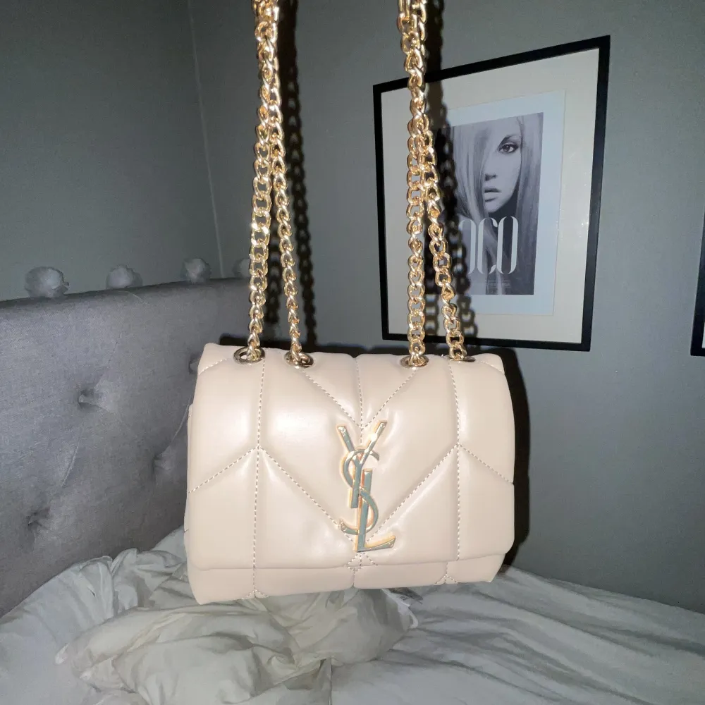 Snygg beige axelväska från Yves Saint Laurent med quiltad design och guldfärgad kedja. Väskan har det ikoniska YSL-emblemet på framsidan och är tillverkad i skinn. Perfekt för att ge en lyxig touch till din outfit.. Laukut & Käsilaukut.