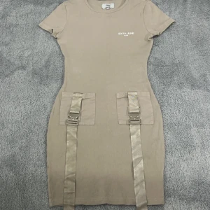 Beige klänning från Sixth June - Trendig beige klänning från Sixth June med korta ärmar och coola detaljer som spännen och band framtill. Perfekt för en edgy look.