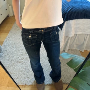 Mörkblå jeans med knappar - Snygga mörkblå jeans med låg midja och bootcut-stil. De har dekorativa knappar på fickorna och en klassisk jeanslook. Perfekta för en avslappnad stil. Gina Young byxor stl 152 passar parfekr för mig som är 152!❣️